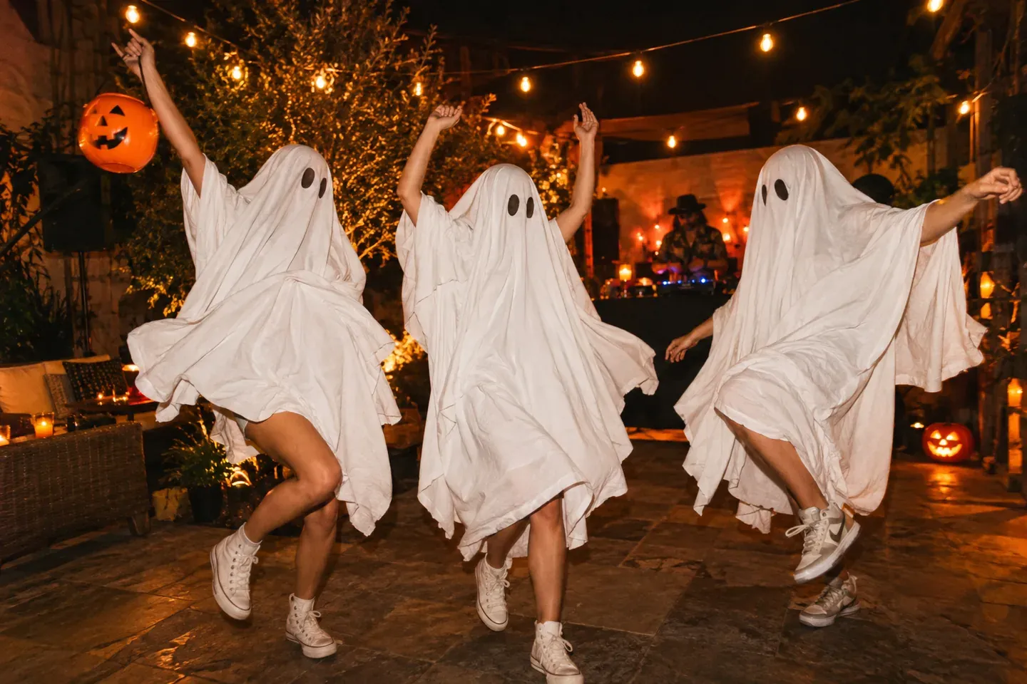 Tres amigos con disfraces de fantasma y tenis bailando en un patio con luces cálidas